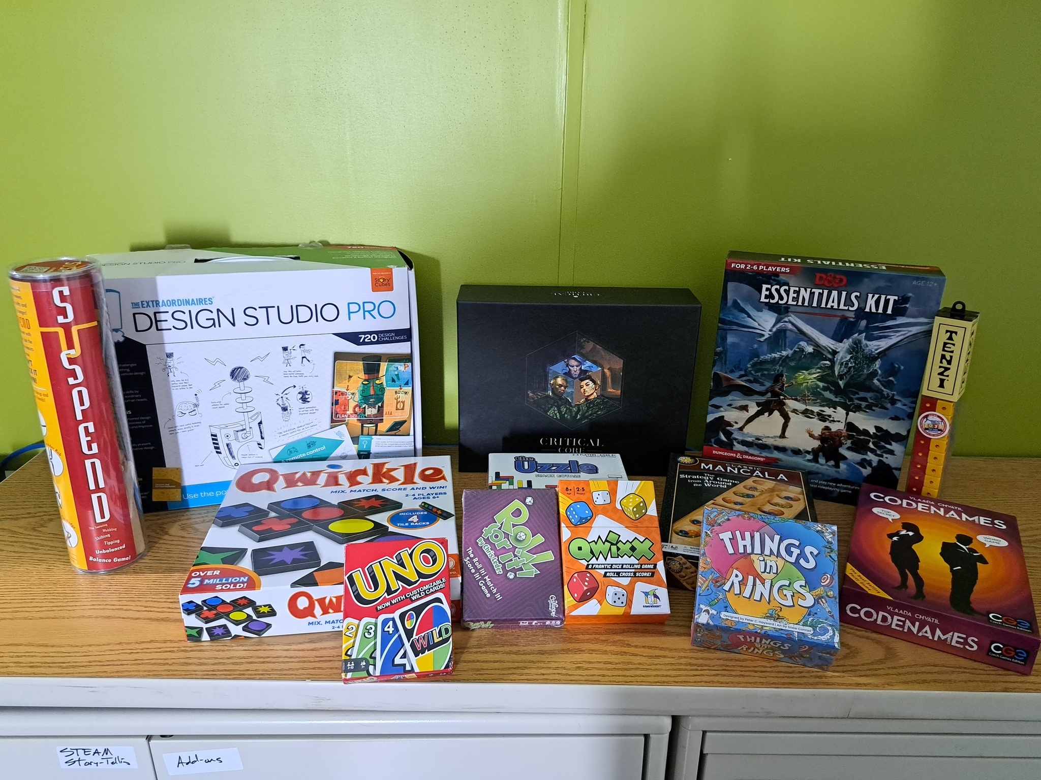 A collection of various tabletop and card games arranged on a wooden surface against a bright green wall. Notable titles include "Codenames," "D&D Essentials Kit," "Qwirkle," "Uno," and "Suspend."
